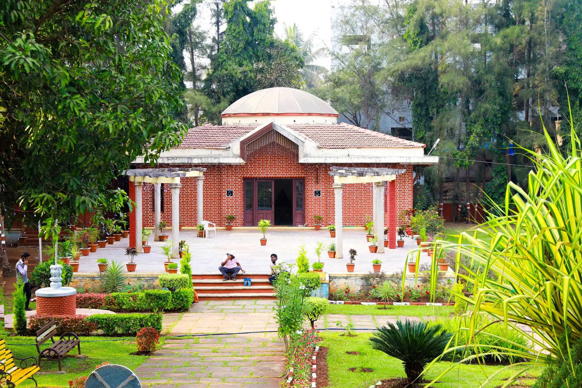 A tiled and red-brick pavilion in a city garden in Belagavi, potted palms along its terrace and people sitting on the steps